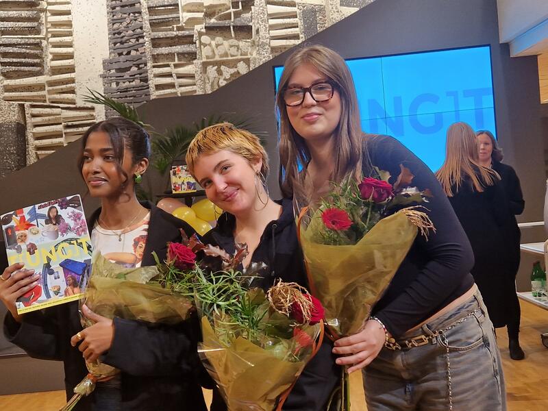 Tre unga kvinnor står med blommor i händerna och ler stolt in i kameran. I bakgrunden av dem syns folk som står och pratar med leenden på läpparna. 